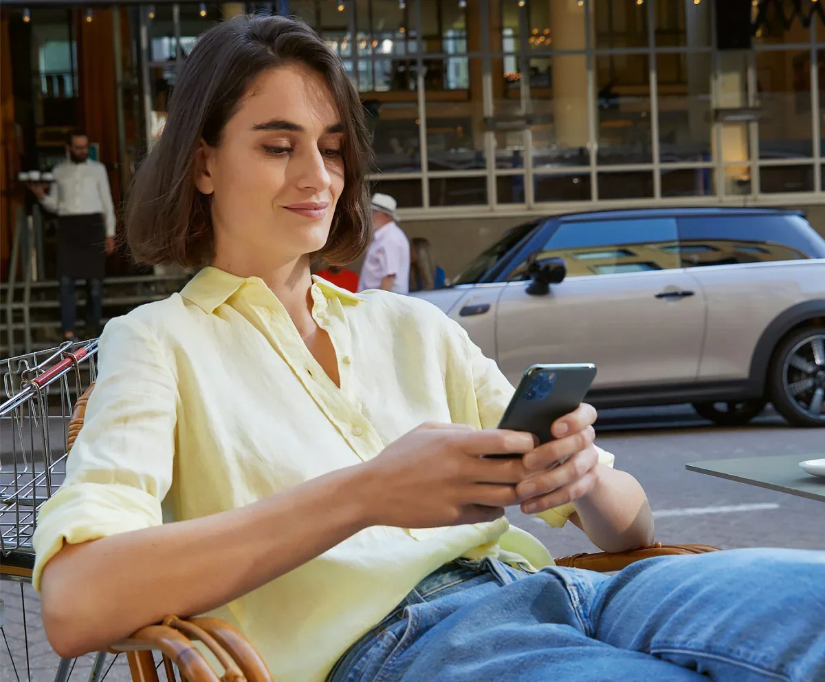 Female wearing yellow collared button-down shirt and jeans, sitting in a chair and holding a smartphone next to a street with a grey MINI vehicle parked in the background. | MINI of Alexandria in Alexandria VA