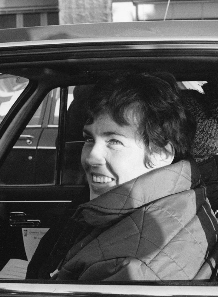 Pat Moss sitting in the driver’s seat of a car, in a black and white photo, smiling and glancing out the window