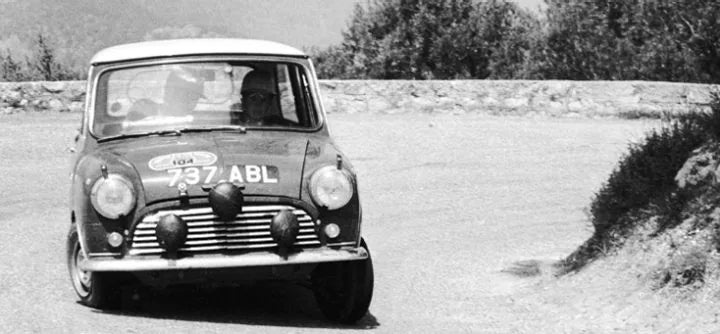 MINI vehicle rounding the corner of a bend, in a black and white photo, with trees in the background.